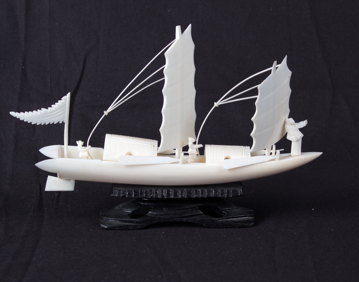 Modell av kinesisk fartøy utskåret i bein på sort sokkel. Skipsmodell med to master og seil, samt tre menn ombord. To par årer langs skutesiden. Vimpel akter.