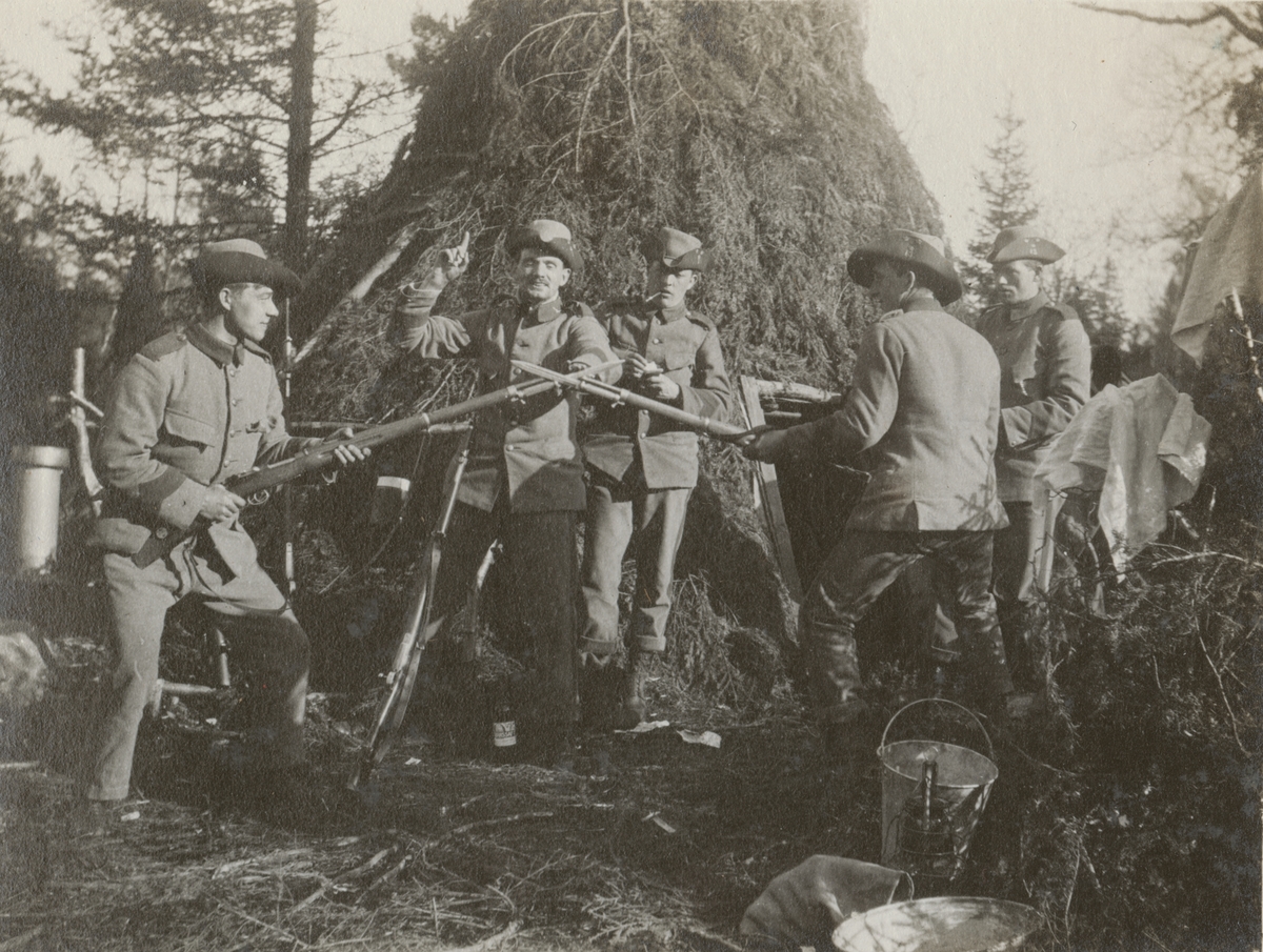 Soldater från Göta livgarde I 2 framför en bivack i skogen. Två av soldaterna är uppställda i närstridsliknande position.