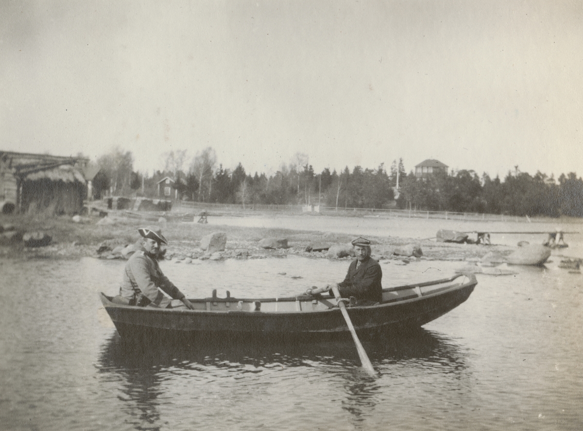 Två män sitter i en roddbåt. Den civillklädde håller åror och soldaten sitter i akter.