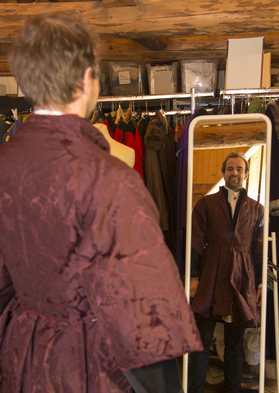Mann i burgunderrød brokadejakke med store skjørt ser på seg selv i speilet og smiler.