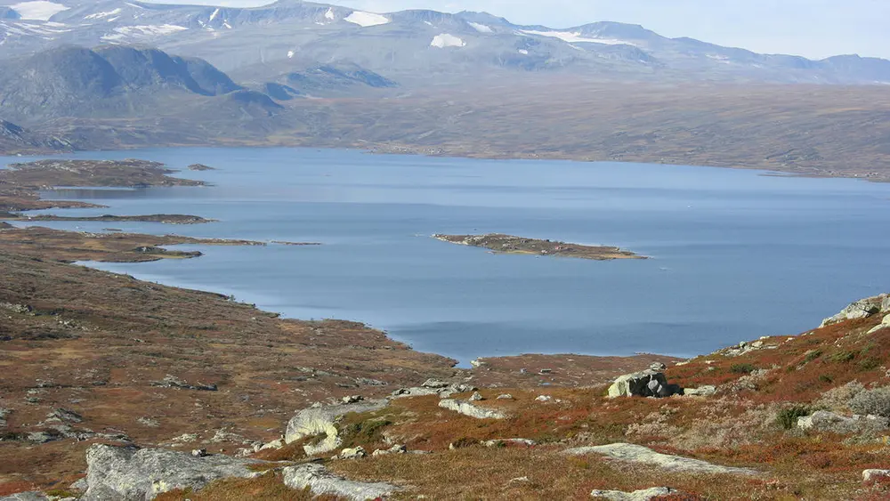 Innsjøen Vinstre sett fra Dragarskaret, i forgrunnen steiner og brunt gras, fjell med litt snø i bakgrunnen.