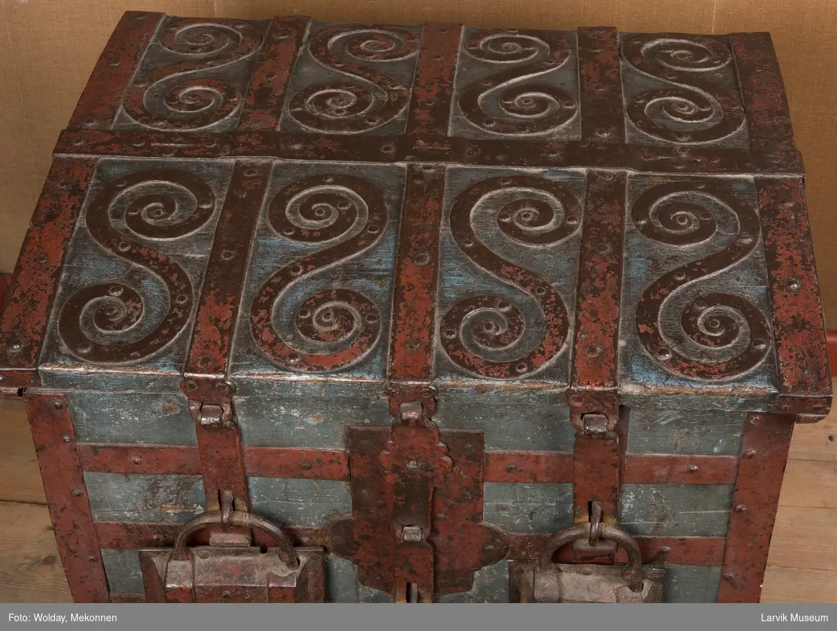Kiste med buet lokk, beslått med rette, krysslagte  jernbånd. 8 dobbeltspiraler av jern på oversiden av kistelokket. 1 lås og 3 hengelåser på ene siden. 1 håndtak på hver kortside.
