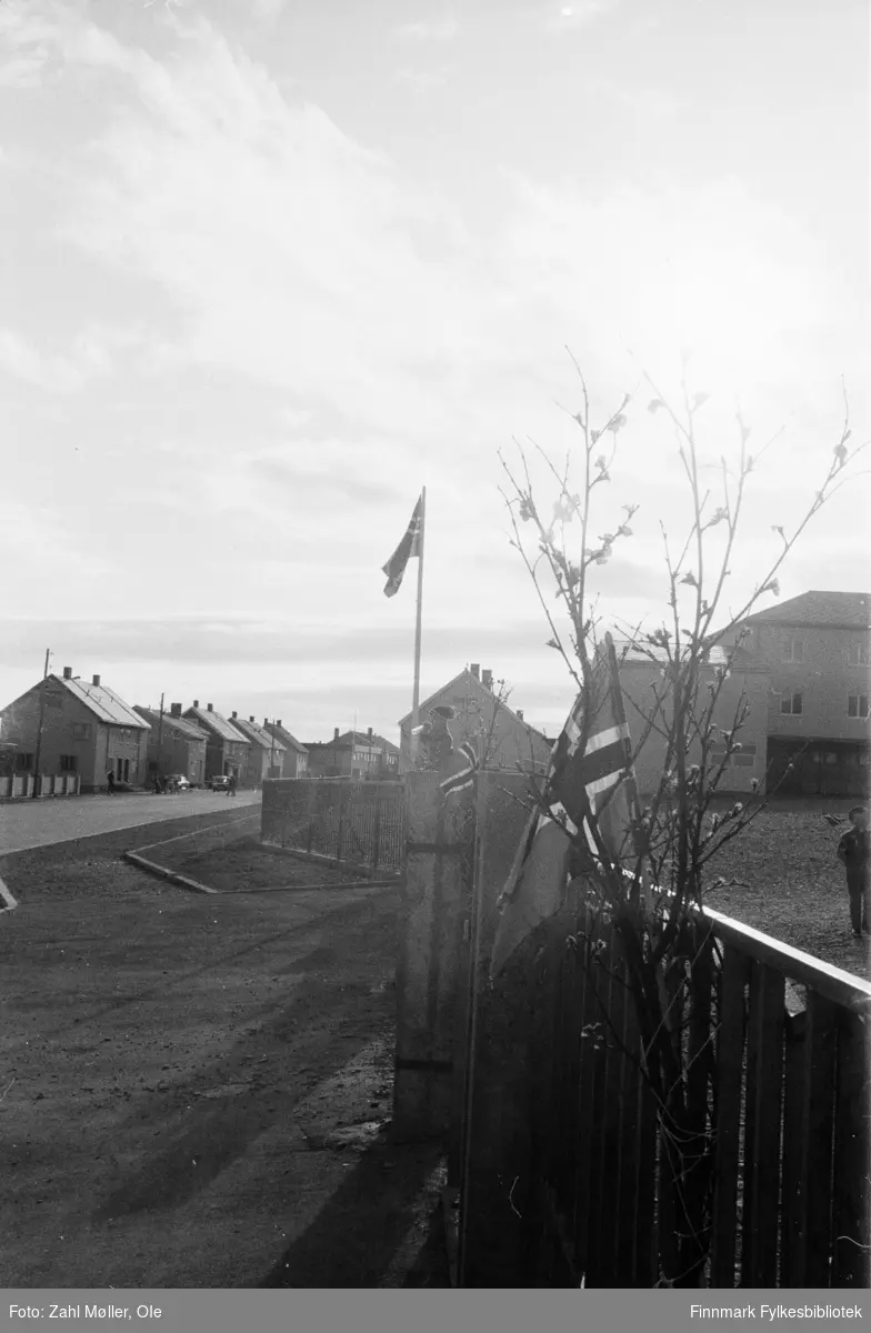 17.mai i Vadsø 1979. Fotografert av Ole Zahl Mölö. Bildet viser detaljer av et gjerde pyntet med flagg og kvister med løvsprett. I bakgrunnen skimtes barn i lek, et barn har klatrert opp på gjerdet eller porten.
