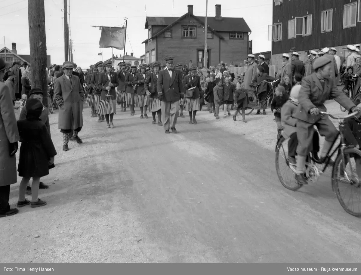 Vadsø 17. mai 1951. Musikkkorps marsjerer i Indrebyen, i Nyborgvegen .Vi ser flagg og fane. Fremst i toget et kvinnelig korps med dirigent. Publikum, barn og voksne sees langs 17.maitoget. Bygningen nærmest til høyre i bildet er "Alders Hvile". Fremst i bildet ser vi en gutt på damesykkel med et mindre barn sittende bakpå sykkelen.