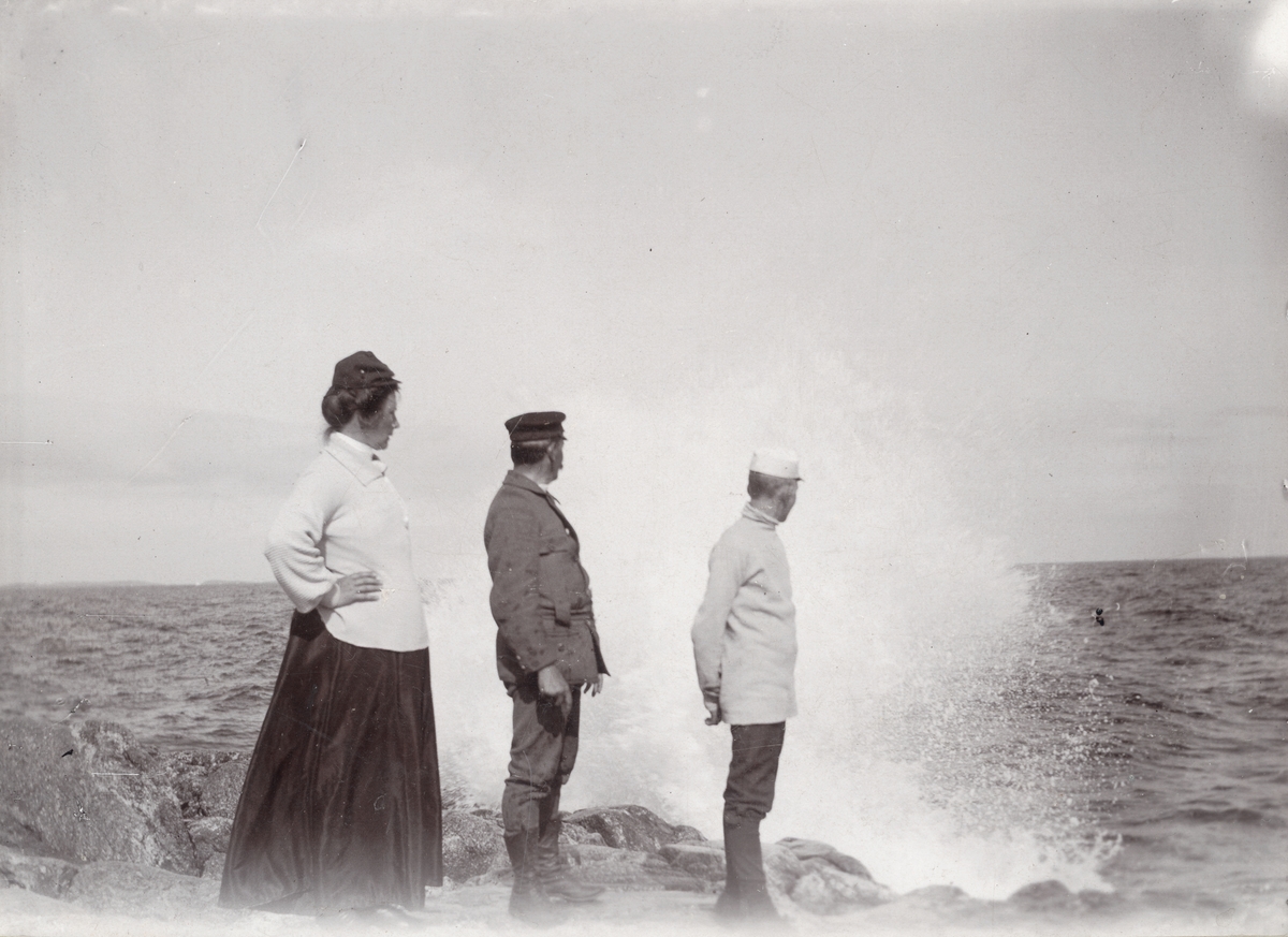 En kvinna och två män på en klippa vid havet, i bakgrunden slår en våg upp. "'Tripp, Trapp, Trull' försjunkna i beundran af hafsscenerier vid Landsort den 24 aug. 1905".