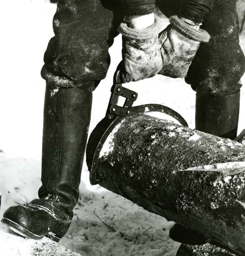 Med handsbeklädda händer lyfts en stock med hjälp av en timmersax. Vintern har täckt marken med ett snötäcke och skogsarbetaren bär kraftiga vinterstövlar av läder som skydd mot kyla och snö.