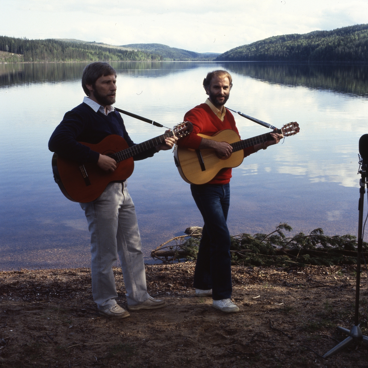 "Afton vid Ängratörn". Två män står vid stranden av en spegelblank sjö och spela gitarr och sjunger.