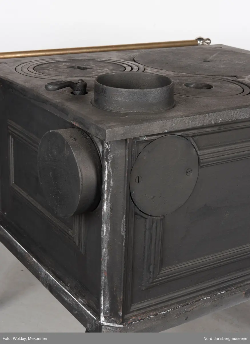 En kvadratisk komfyr med tre dører, tre kokeplater med ringer, og åpning for varmtvannbeholder. Røykrør  opp fra topplate, høyre bakside, med spjeldregulator. Messingfarget håndklestang i front.
