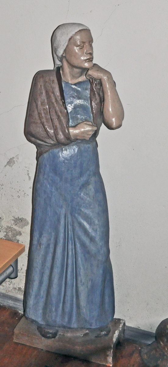 Maria-figur - står på gulvet, ser mot krusifikset på veggen.