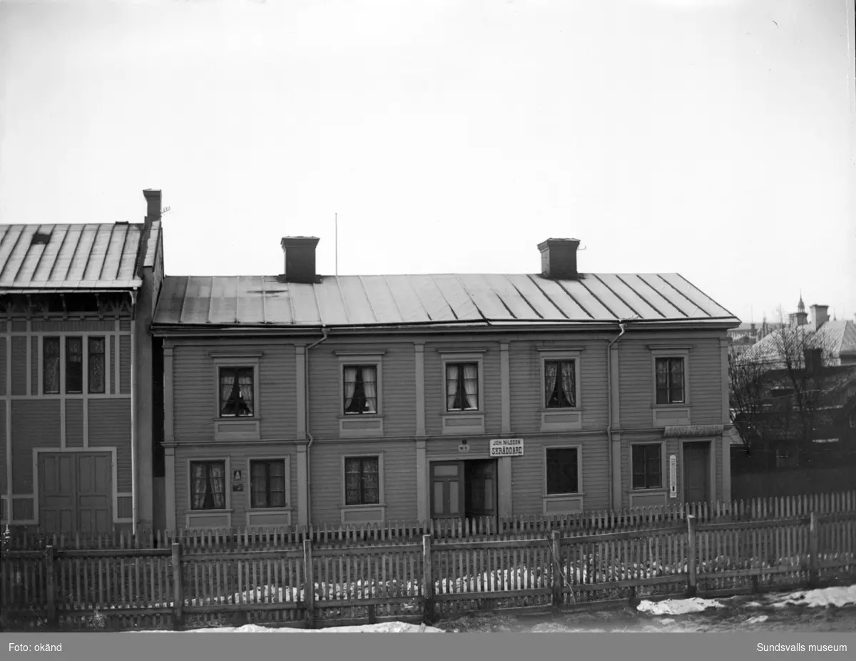 Bostadshus efter Norrmalmsgatan 7-9. Skyltar på fasaden: "Joh. Nilsson SKRÄDDARE" samt Ernst Westman URMAKARE".