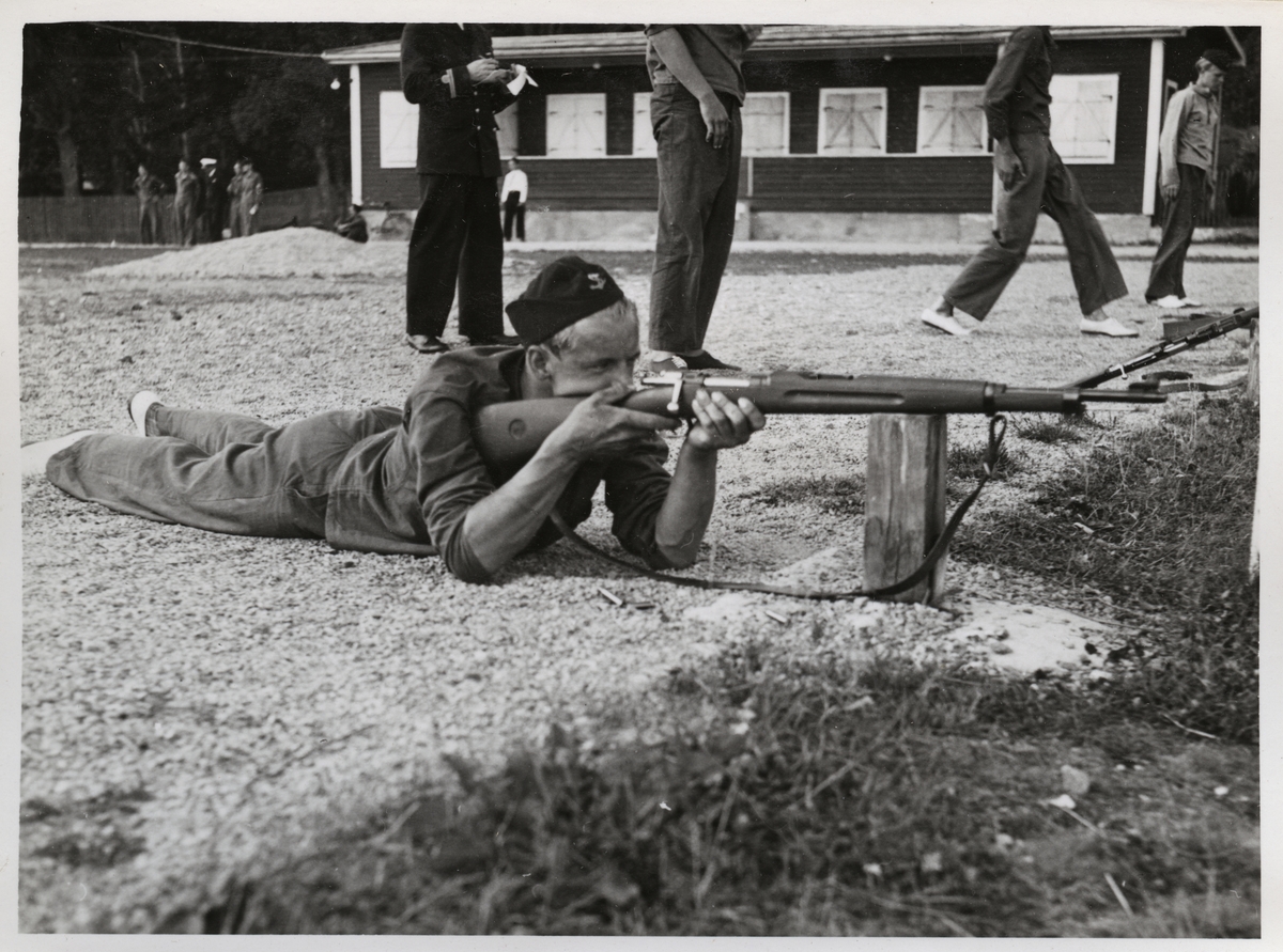 Bilden visar en rekryt under skjututbildning med gevär. Han ligger på mage och siktar mot målet. I bakgrunden syns en officersom antecknar i ett block och flera värnpliktiga.
