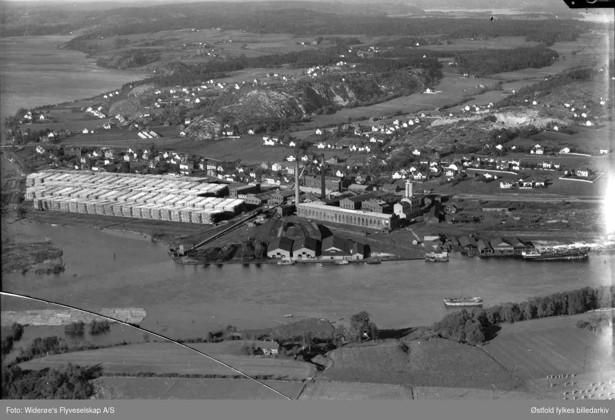 Greåker Cellulosefabrikk i Tune med Glomma, stabler med tømmer eller plank.
Bebyggelsen og  Visterflo ses ibakgrunnen. Flyfoto 1935- 40 ca.