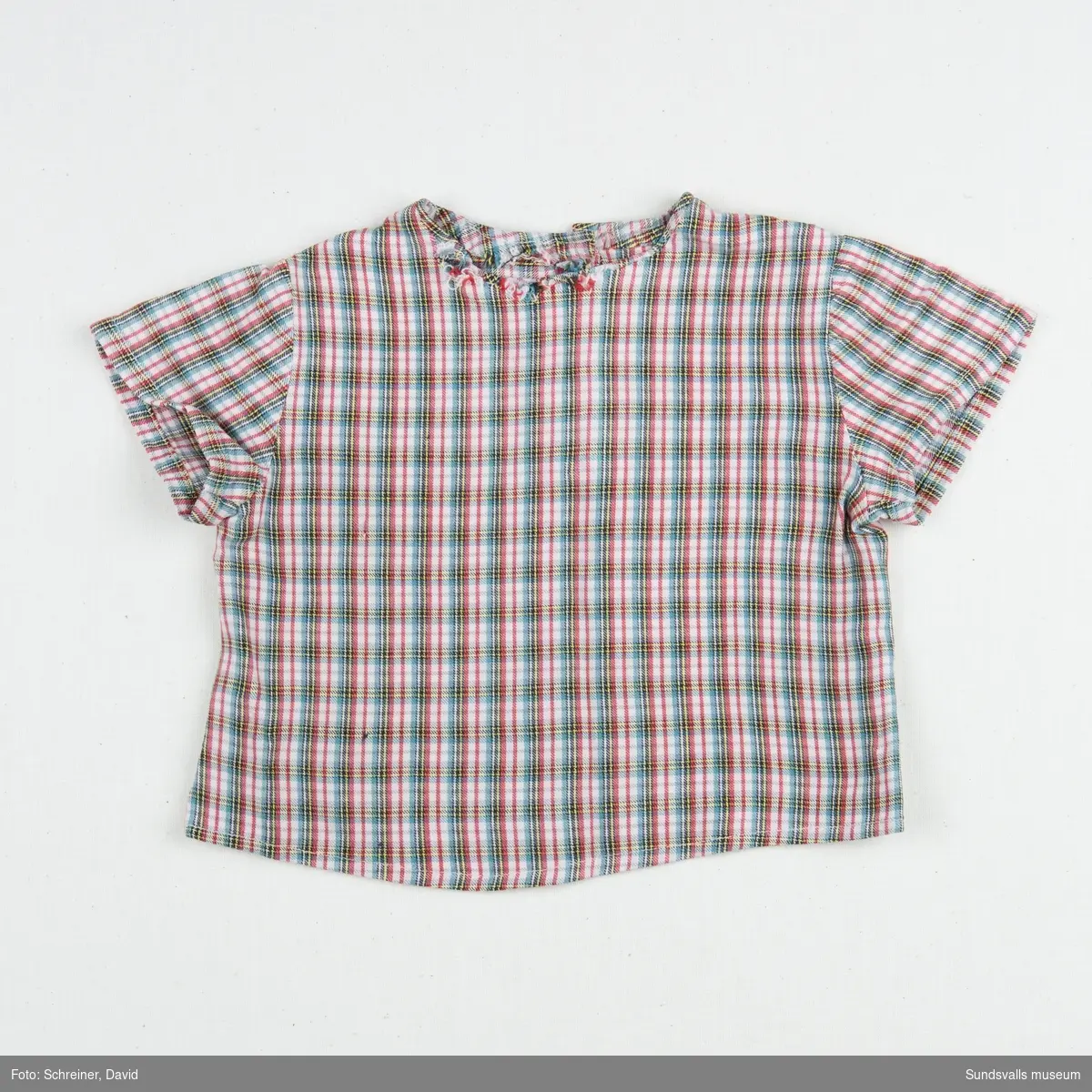 En kortärmad smårutig pojkskjorta med rundad hals som knäpps med två vita plastknappar i ryggen.