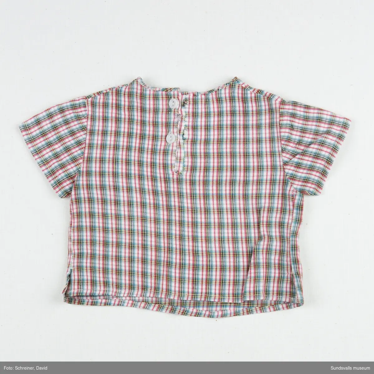 En kortärmad smårutig pojkskjorta med rundad hals som knäpps med två vita plastknappar i ryggen.