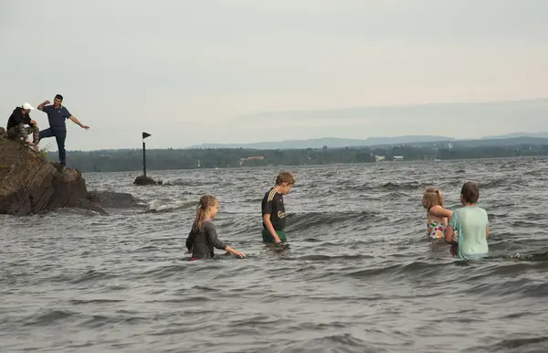 Four young people have gone swimming in Lake Mjøsa with their clothes on, and on a cliff two more people are standing and watching.