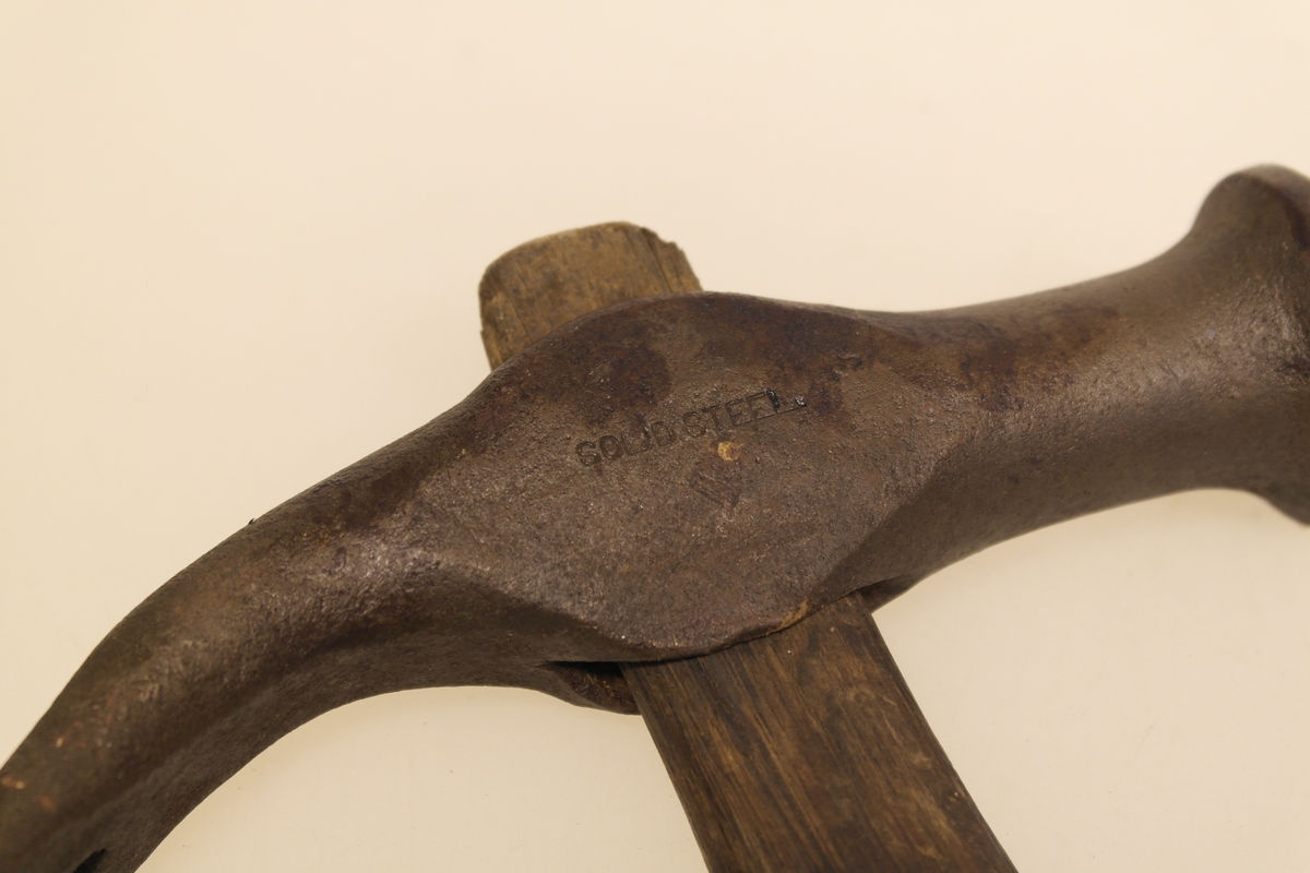 Skomakerhammer i jern, med skaft av skåret tre. Skaftet er tappet gjennom hodet. Stempel/Innskrift på jernet.
