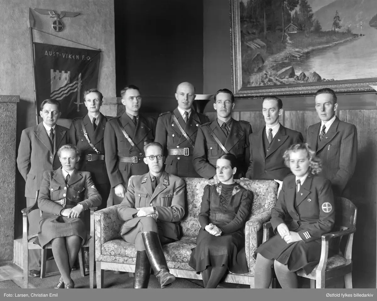 Nasjonal Samling i Sarpsborg, fane med tekst: Aust-Viken F.O.Nasjonal Samling (Fylkesorganiasasjon)
Uniforimert mann i midten, fylkesfører, Olaf Hoff. 
Damen lengst til venstre foran har armmerke på uniformen: Romerike. Ørn og solkors på veggen. 
Solkors, ørn og tre stjerner - armbind på fylkesfører.