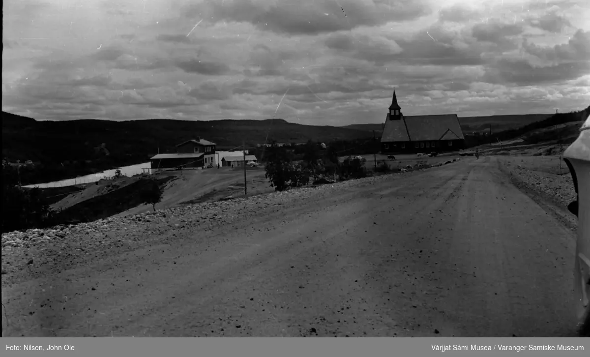 Masi kirke og et hus med et vann i bakgrunnen. Kyr beiter foran kirka. Máze / Masi, Juli 1967.
