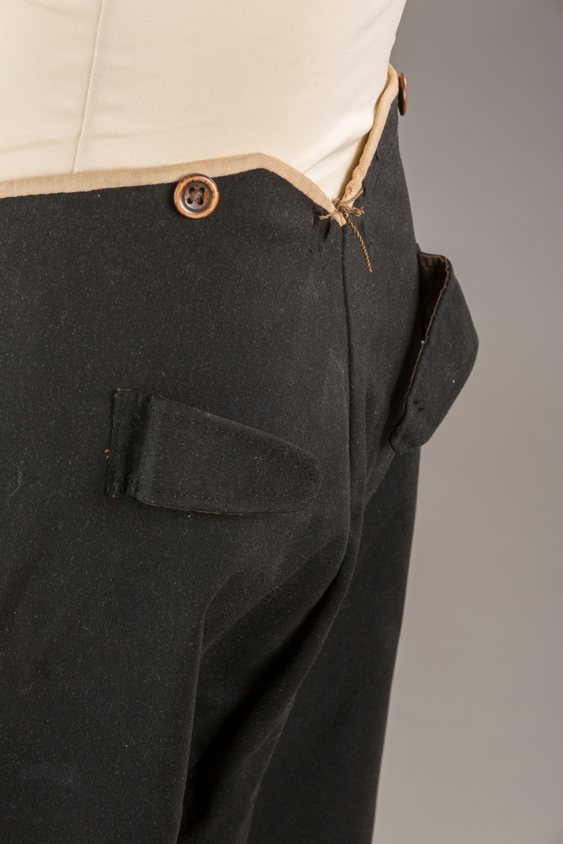 Form: Bena utvidet nederst. Lerretskant rundt livet, spensel i ryggen, mangler spenne i sepnsel?
Delvis foret. To lommer. 5 knapper, mangler en knapp.