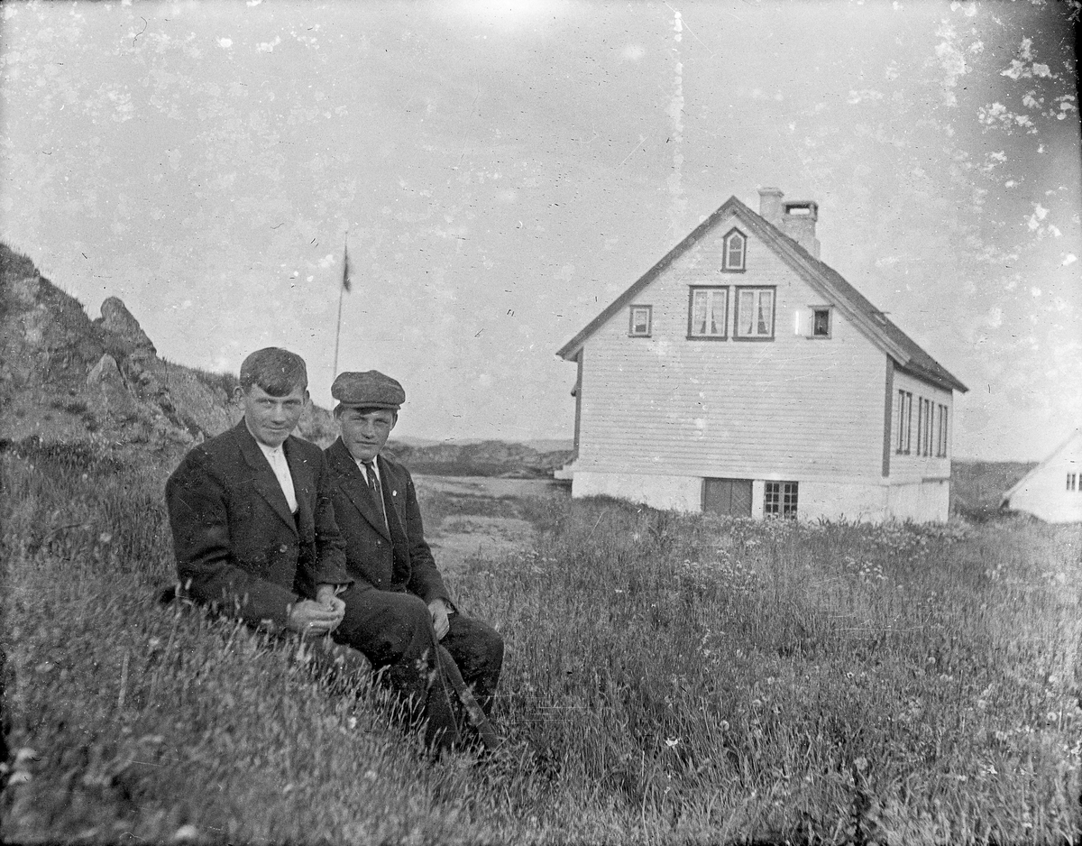 Gruppebilde. 2 unge menn sittende i gresset med bolighus i bakgrunnen. Flagget er oppe.