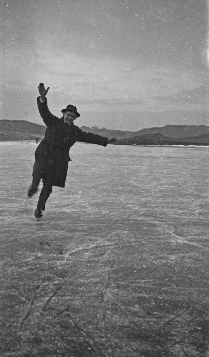 En mann i hatt og frakk viser sine ferdigheter på isen. Skøytene er av typen man fester på vanlige sko. Hverdagsfoto.