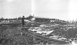 Utgraving. Byggeplass. En mann i hatt og frakk står og ser p