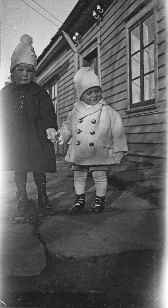 Barn. To småjenter holder hverandre i hendene på hellene foran inngangsdøren til en bygning. Hverdagsfoto.