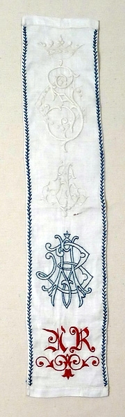 Märkningsprov sydda på vitt linne i vitt rött och blått. Överst en krona och två monogram i vitt, därunder ett monogram i vitt och blått och längst ner ett i rött med bladslinga under. Maskinfållat och över de två längsgående sömmarna kråkspark i blått.