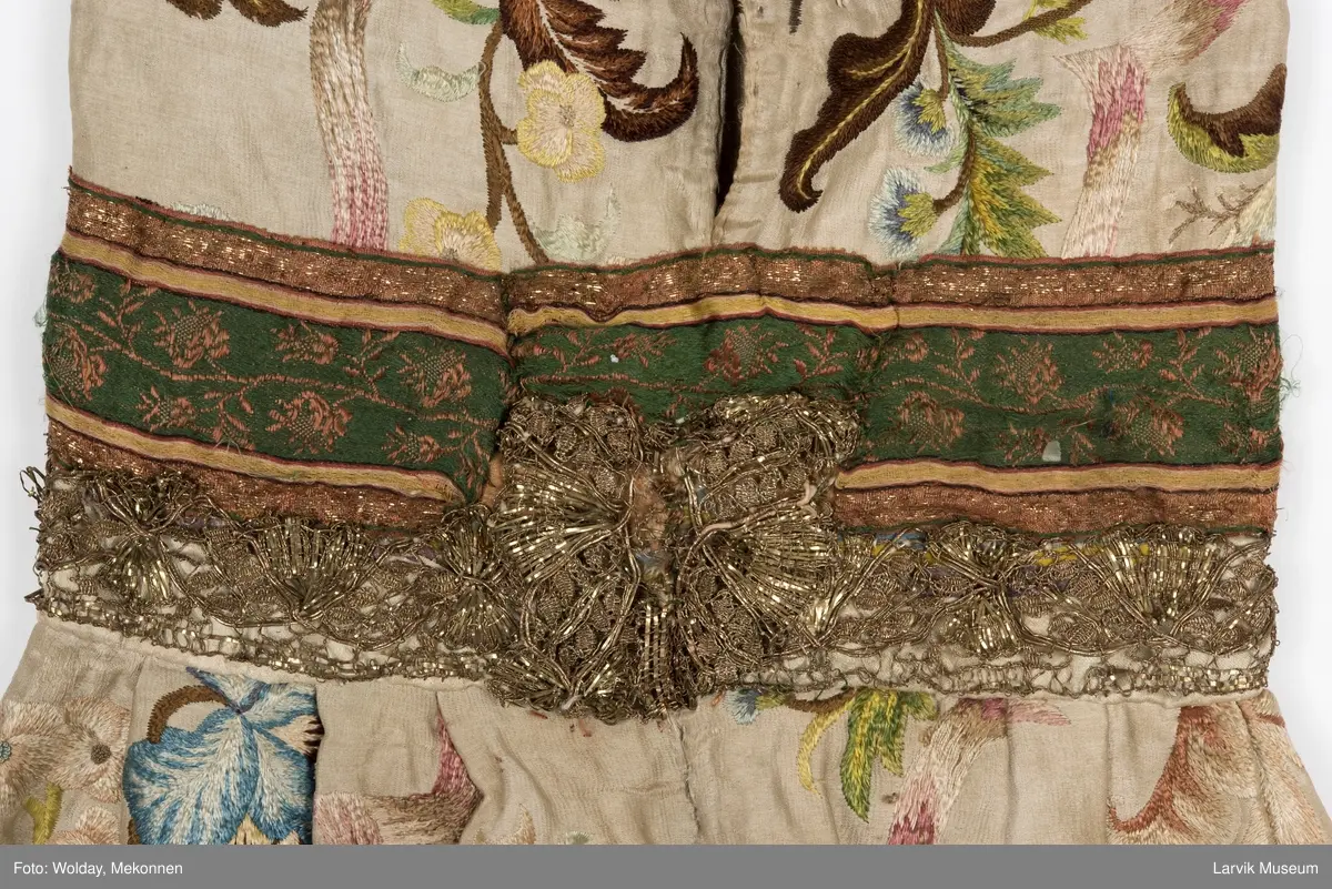 Form: livstykke som hette, åpen til livet, bred linning, rynket skjørt med blondekant nederst, søm bak på hetten i linning, søm foran og bak i skjørt, enkelte skjøtesømmer, linning brodert med gulltråd, skjørtet er foret med tynt stoff