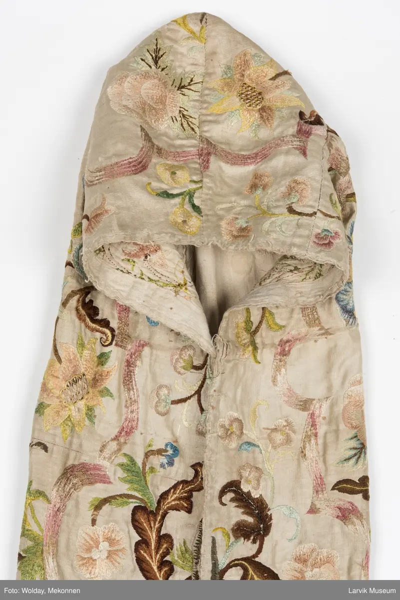 Form: livstykke som hette, åpen til livet, bred linning, rynket skjørt med blondekant nederst, søm bak på hetten i linning, søm foran og bak i skjørt, enkelte skjøtesømmer, linning brodert med gulltråd, skjørtet er foret med tynt stoff