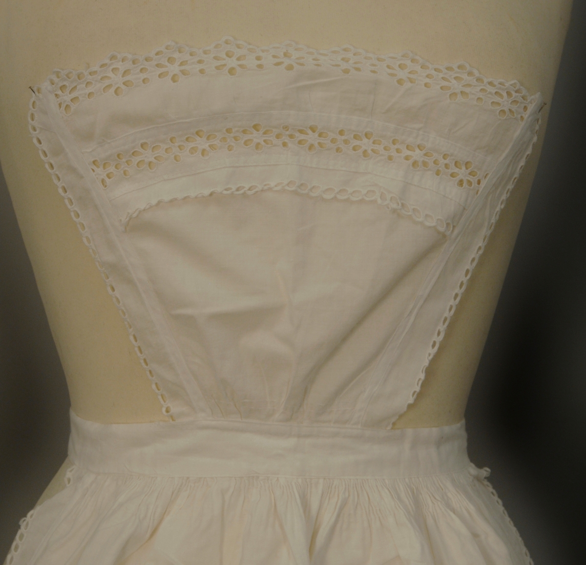 Kvitt (serverings)forkle av staut (lerretsvevd bomullstøy) med blonder. Nedentil er forkleet påsydd en engelsk blonde (4 cm), ovenfor er et mellomverk (2,5 cm) og over der igjen en rad med engelsk blonde og over der en ca 2,5 cm bred remse med stoff med små hull som vender oppover. Samme enkelthullete blonde oppover langs begge sidene. Øverst en liten "lapp" hvor det er samme blondeoppsett som nederst. Forkleet har bånd til å feste rundt livet. Blondene på høyre side (sett forfra) er slitt nederst og litt lengre opp.