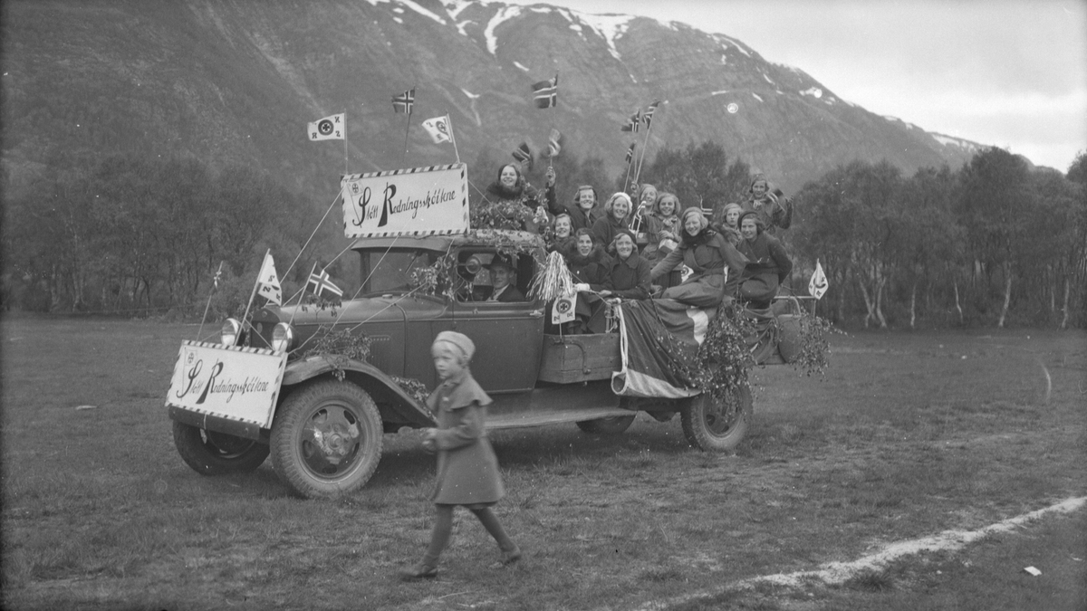 Bil dekorert med flagg og bannere, Støtt Redningsskøytene, masse ungdom på lasteplanet.
Aksjon til støtte for Redningsselskapet. Bilen er en Ford AA 1930-31.
