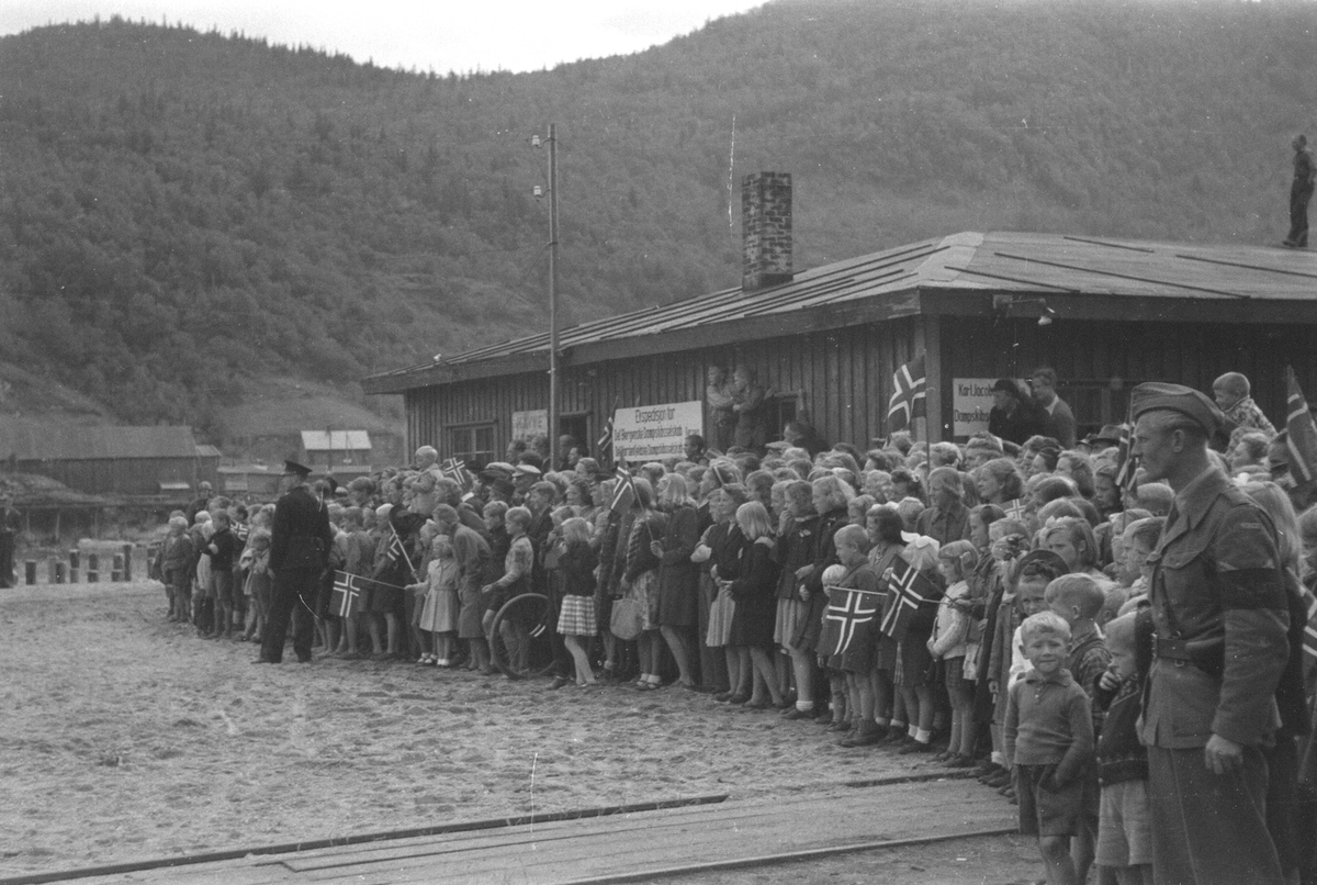 Kronprins Olav ankommer Mosjøen etter frigjøringen. Stor folkemengde venter på Kronprinsen ved den gamle dampskipsekspedisjonen og havnekafeen.