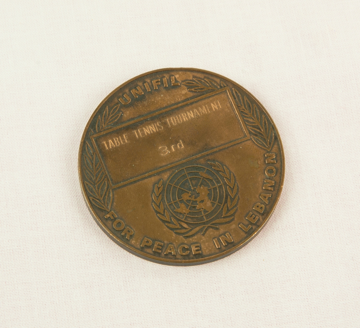 Medalj i metall från mästerskapen i bordtennis från FN-insatsen i Libanon. Medaljen är för tredje plast och är gjord i brons. På ena sidan av medaljen finns en FN logga omgiven av en lagerkrans, under står "UNIFIL FOR PEACE IN LEBANON". På andra sidan finns en karta över Libanons västra del samt delar av Israel och Syrien. Medaljen förvaras i ett lackat träfodral som även fungerar som stöd. Fodralet har en filtklädd insida.