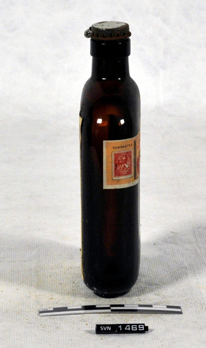 Brun flaske med kork og etikett.
Korken er festet til flasken med hengsel i metall.
Flaskens form er en tynn oval
