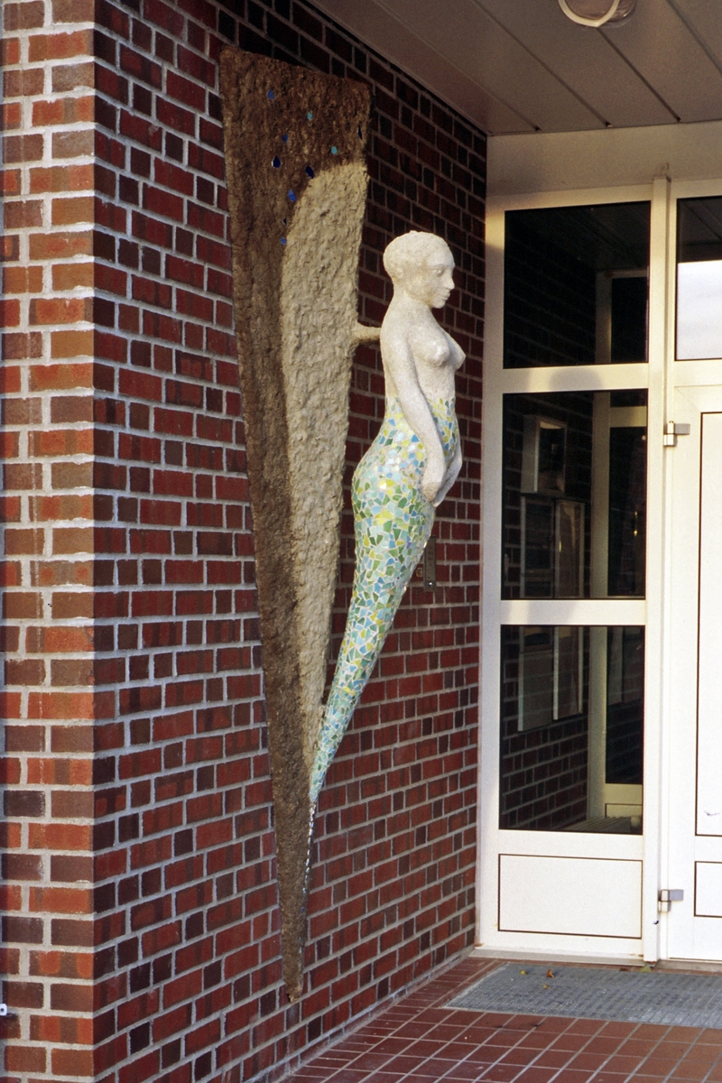 Utsmykkingen består av en figurativ veggskulptur utformet som en kvinneskikkelse/gallionsfigur. Figuren fremtrer på en bakgrunnsform, et relieff som gir assosiasjoner til baugen på et skip.