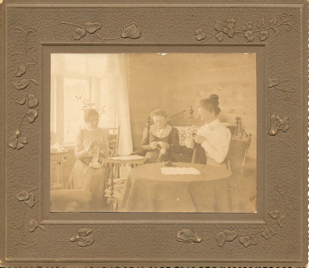 Gruppe kvinner. Tre kvinner sitter med håndarbeid (broderi eller hekling) ved vindu. Nanna Pedersen, fru Laws, frk. Aabakken. Interiør. Umalte plankevegger.