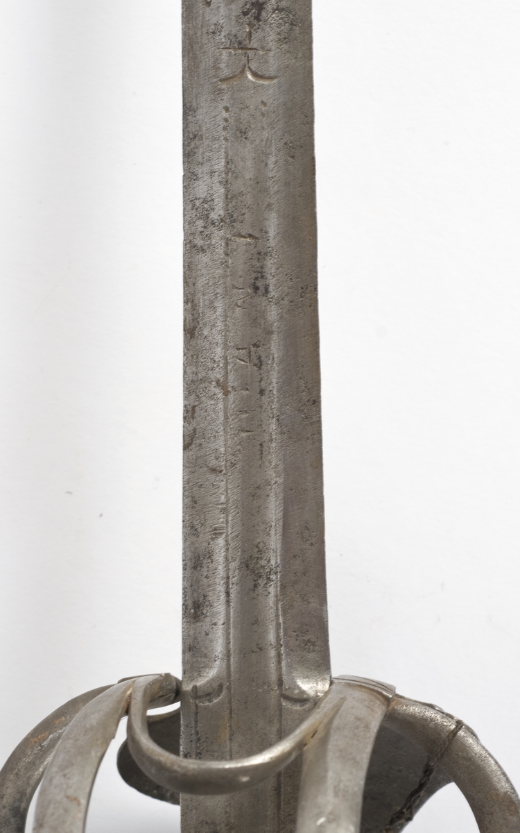 Plommeformet  8 kantet knapp. Tregrep.. Asymmetrisk feste med flate parerbøyler.  S formede parerstenger..Fremre parerstang går over i håndbøylen.Oppbøyet skjellformet parerplate på innsiden.
Tveegget, rett klinge med 6 kantet tverrsnitt  Kort blodrand merket IVAN