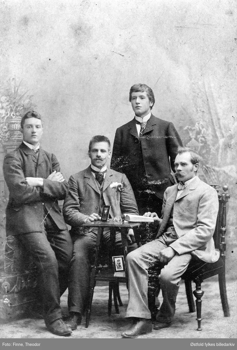 Gruppeportrett tatt i atelier i kabinettformat av fire ukjente menn, to stående og to sittende. Dett er et iscenesatt portrett med fire ukjente menn ved et bord med utstilte fotografier, en leser i bok. Denne type iscenesatte motiver var populære rundt 1900.