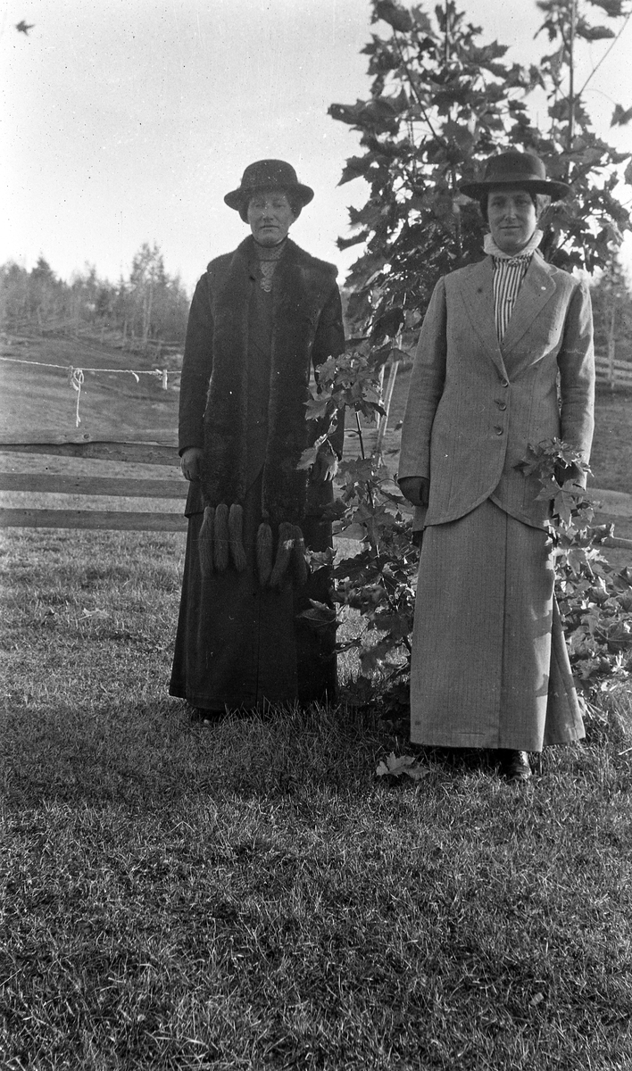 To kvinner i helfigur ved et tre. De to er Marie Lund, f.Simensen (til venstre) og søstera Borghild Elise Woldheim, begge bosatt i Gjøvik, men bildet kan være tatt ved bruket Nordeng i Øverskreien.