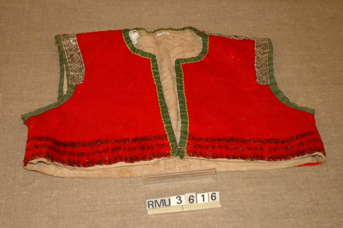 Rødt livstykke i ull med grønne og gulstripete kanteband i bomull. Livet er også dekorert med metallband. Merker etter isydde ermer.
