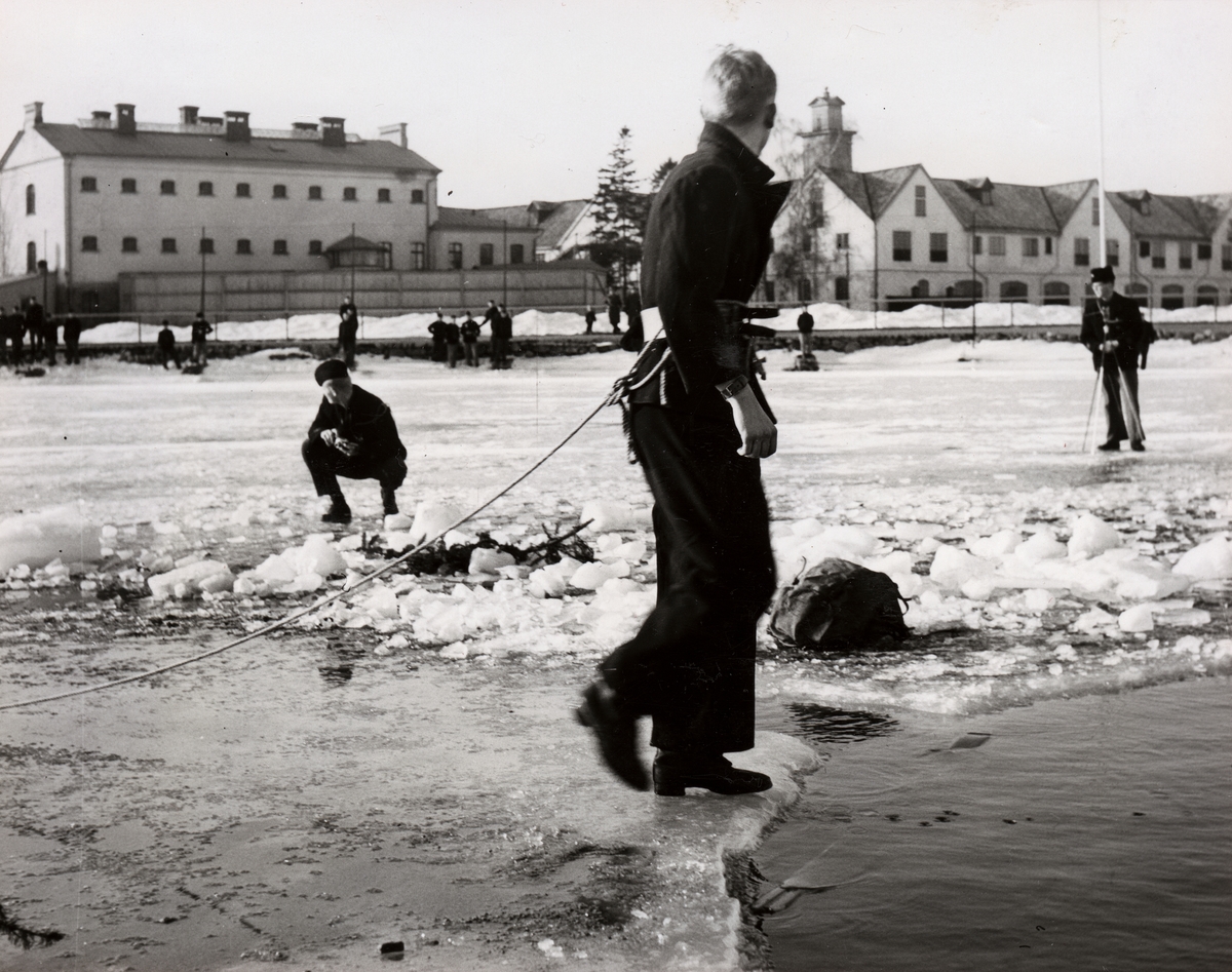 Man som står vid en isvak med rep runt midjan övar på livräddning. I bakgrunden syns kronohäktet och slup- och barkarskjulet.