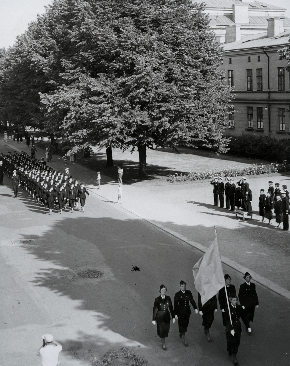 Uniformsklädda lottor marscherar längs med Vallgatan utanför Bataljon af Trolle i Karlskrona. Utanför bataljonen står bl.a. en konteramiral och gör honör.