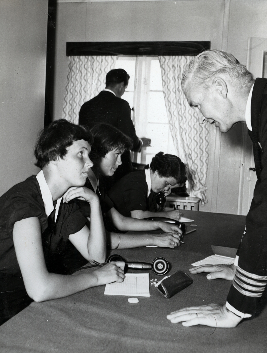 Tre kvinnor sitter vid ett bord med telefoner och anteckningsblock framför sig. En uniformsklädd militär av hög grad lutar sig fram över bordet framför den första kvinnan. I bakgrunden står en man vid ett fönster med ryggen mot kameran.