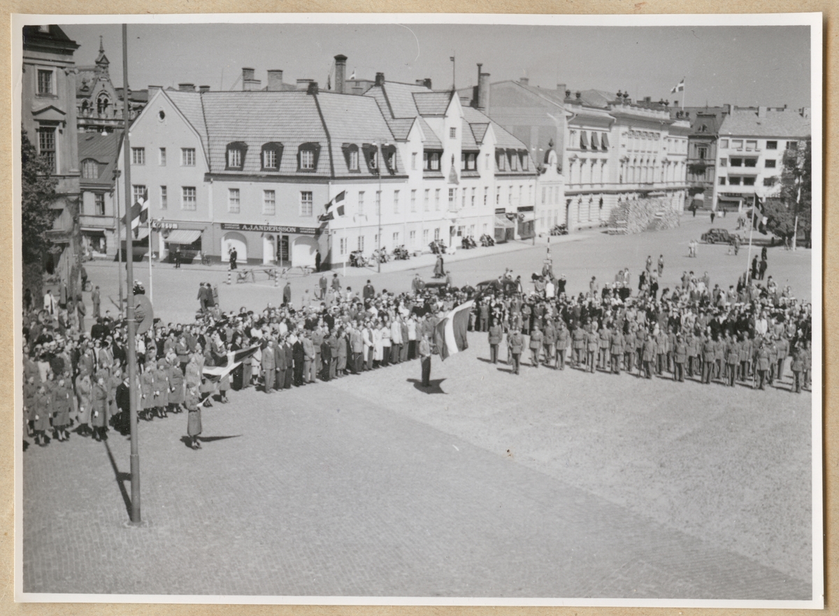 Soldater, lottor och civilklädda människor uppställda på stortorget i Karlskrona. Längst fram står två personer som håller i varsin svensk flagga. I bakgrunden syns kringliggande bebyggelse, bl.a. Karlskrona stadshotell.