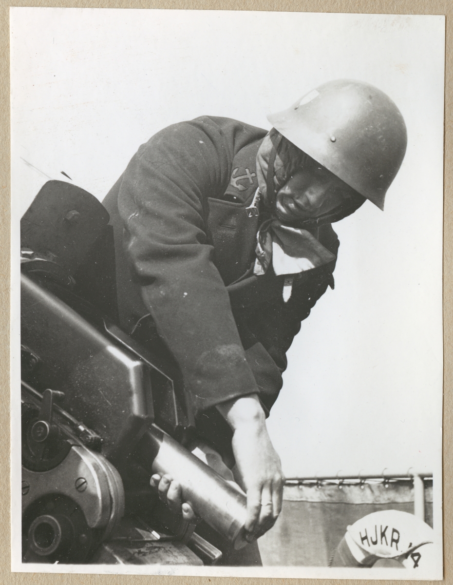 En militär, klädd i uniformsrock och hjälm, lutar sig fram och laddar en kanon med ammunition ombord på hjälpkryssaren Wiros.