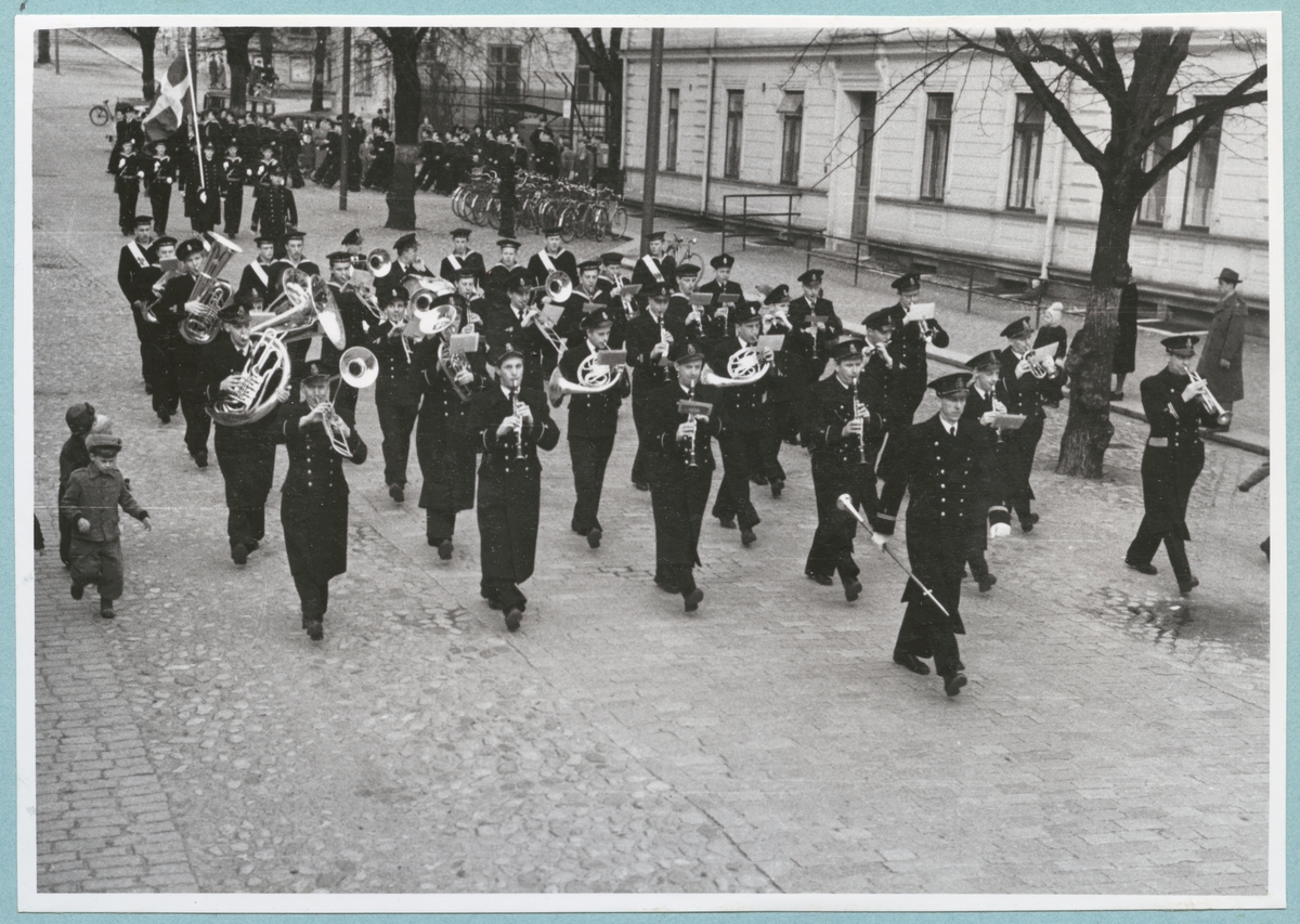 En militär musikkår marscherar längs med Drottninggatan. Ut från bataljon Sparres kaserngård marscherar även flera uniformsklädda flottister ut på gatan. Bredvid musikkåren springer små barn. 
Kyrkparad för nyinryckta värnpliktiga.