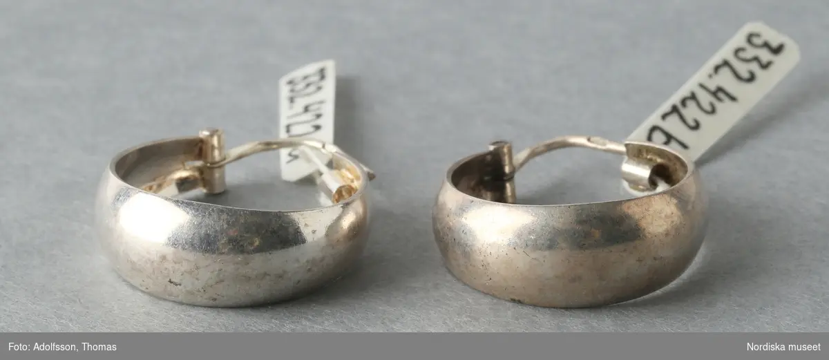 Ett par örhängen, av silver. Breda ringar med låsanordning bestående av smal böjd ten, att fästa genom örat, upptill. Tenen stämplad "925". 
/Leif Wallin 2015-06-08