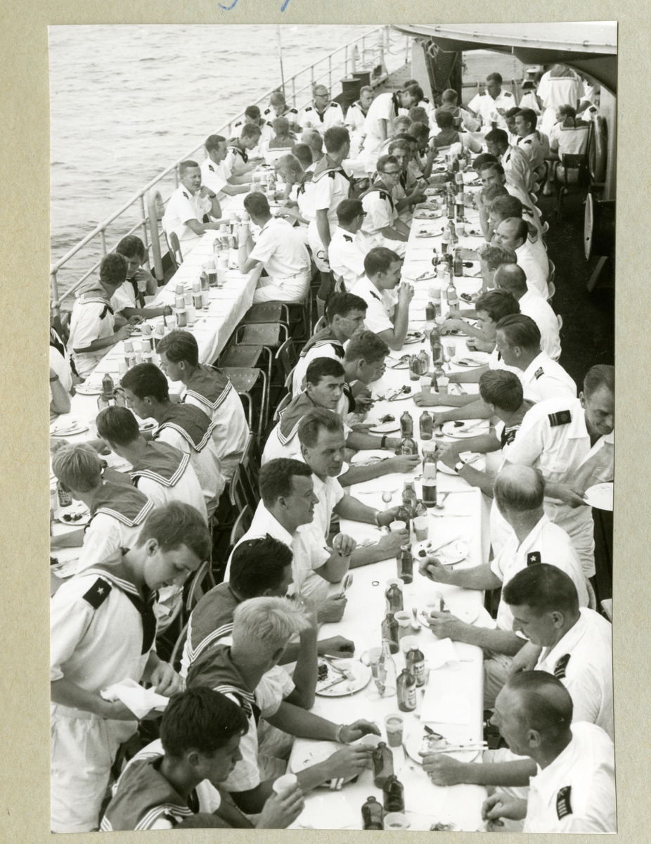 Bilden föreställer två uppdukade långbord på minfartyget Älvsnabbens däck, vid vilka det hålls en middag. Besättningen sitter till bords i sommaruniform. Bilden är tagen under långresan 1966-1967.
