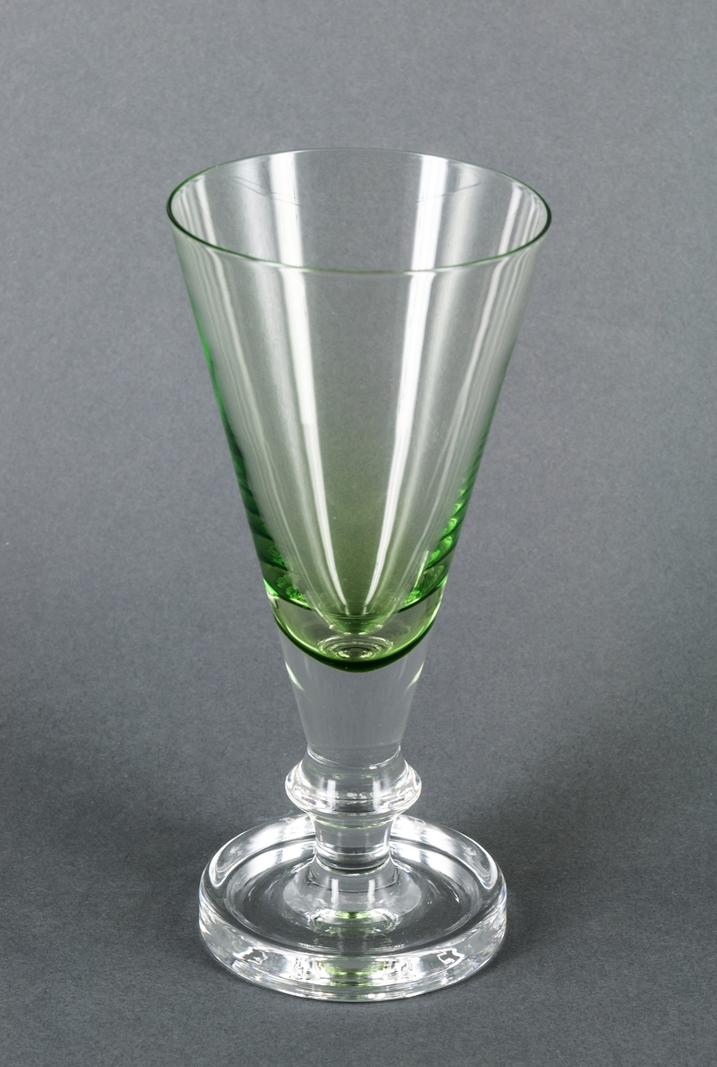 Ölglas i halvkristall med grönt underfärg. Prov. Sandviks glasbruk, design Gunnar Cyrén. Konformad kupa, via utdragning övergående i lågt konkavt ben. Fot med konkav översida, tjockast i ytterkanten.